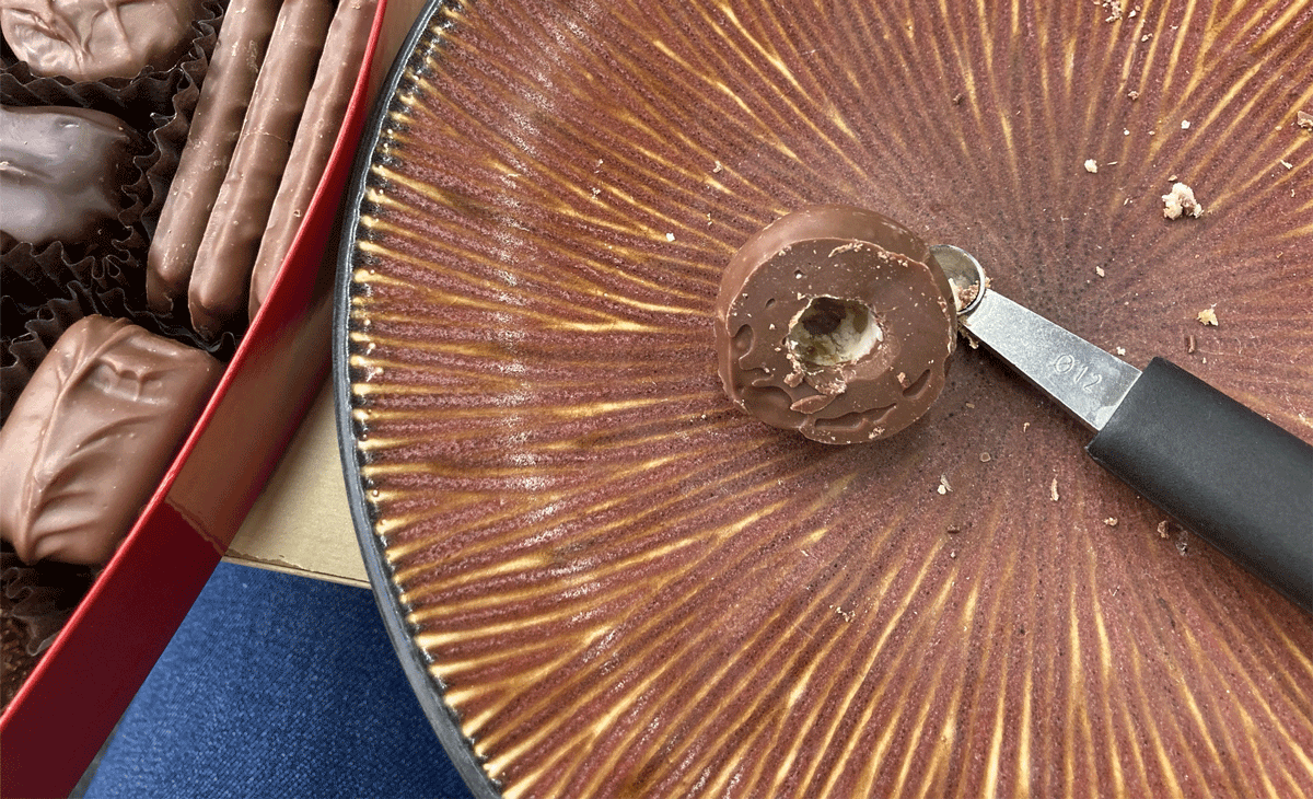 A chocolate candy, with a portion of the bottom carved out, sitting on a plate, alongside a melon baller. 